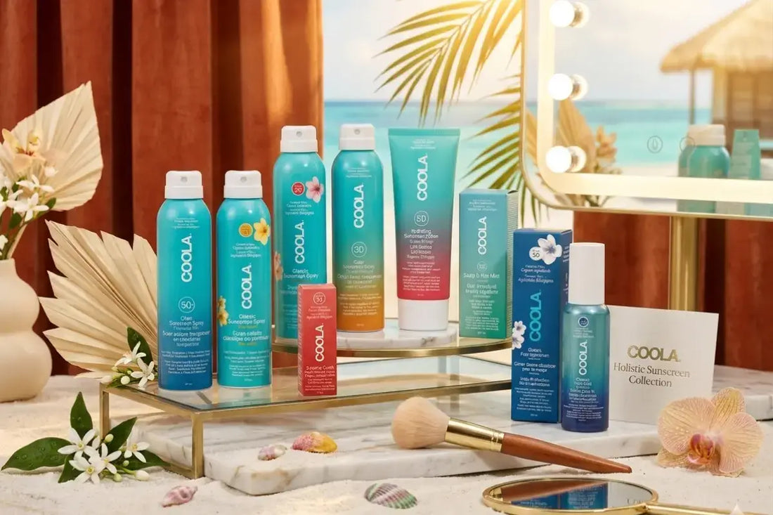 COOLA solkräm-sortiment uppställt på strandtema: sprayflaskor, ansiktskräm och läppbalsam med SPF, omgivet av sand och tropiska blad